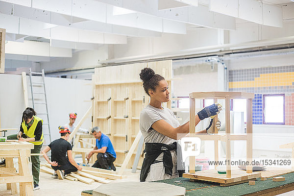 Zuversichtliche junge Tischlerin reibt in beleuchteter Werkstatt Sandpapier auf Holztisch an Werkbank