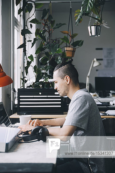 Side view of confident male hipster using laptop at desk in creative office
