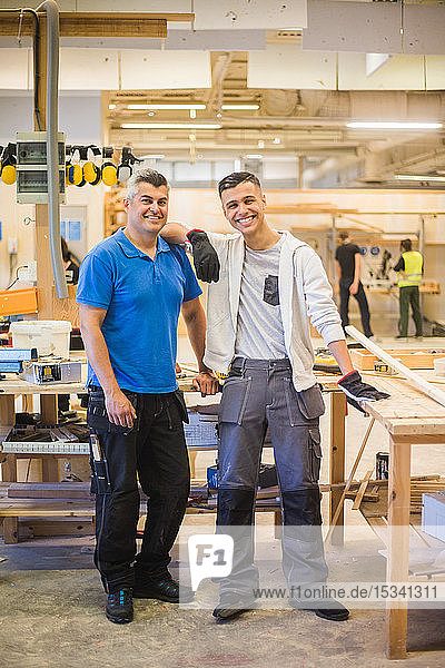 Full length portrait of smiling teenage trainee standing with hand on instructor's shoulder by workbench at illuminated