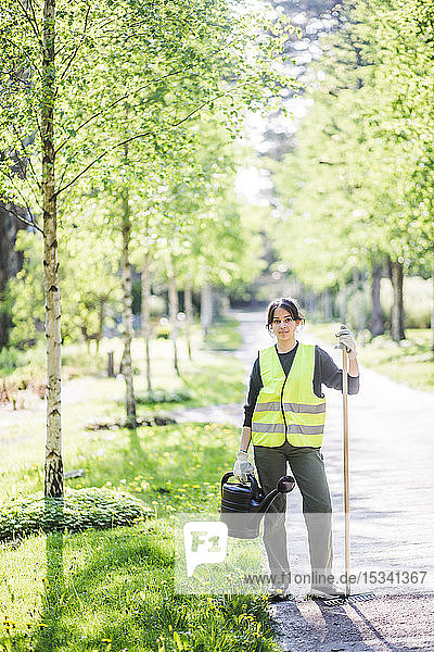 Full length portrait of confident young woman holding watering can and rake on footpath at garden