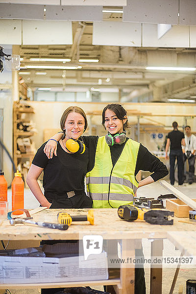 Portrait of smiling young female coworkers standing with arm around at workbench