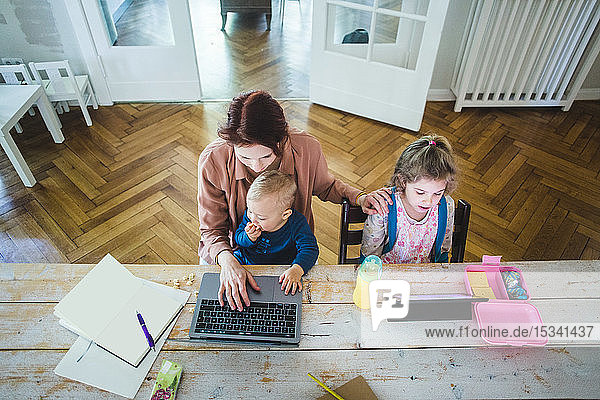 Hochwinkelansicht einer Mutter  die am Laptop arbeitet  während sie mit ihrem süßen Sohn zusammen mit ihrer Tochter am Tisch sitzt und ein digitales Tablett benutzt