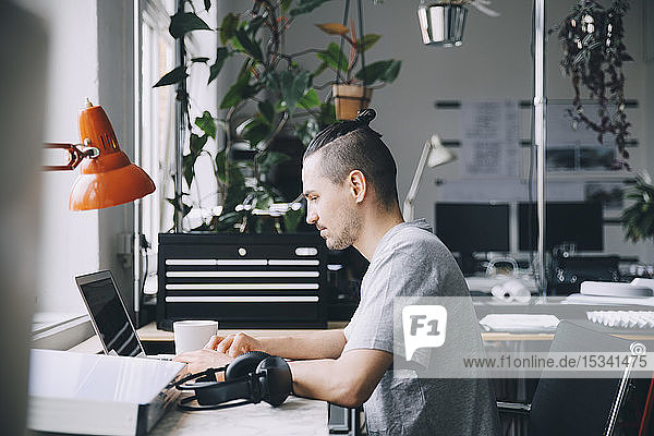 Seitenansicht eines männlichen Hipsters mit Laptop am Schreibtisch im Kreativbüro