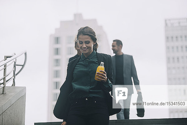 Lächelnde Geschäftsfrau hält Getränk in der Hand  während Kollegen im Hintergrund in der U-Bahn