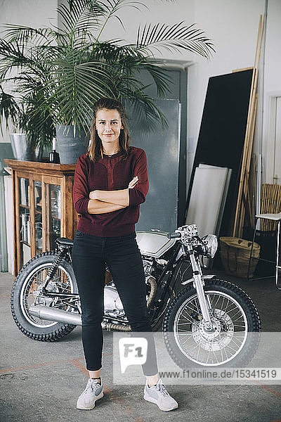 Portrait of confident female entrepreneur standing with arms crossed against motorcycle in creative office