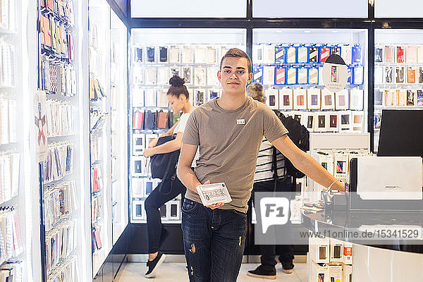 Porträt eines selbstbewussten jungen Verkäufers  der eine Telefonhülle hält  während er mit Kunden im Hintergrund an der Kasse steht