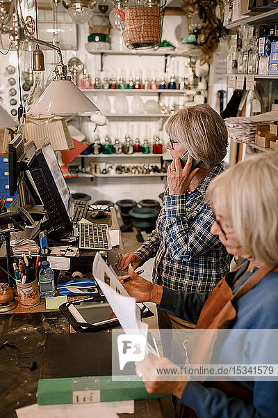 Ältere Eigentümerin liest Dokumente  indem ein Mitarbeiter an der Kasse im Laden mit dem Handy telefoniert