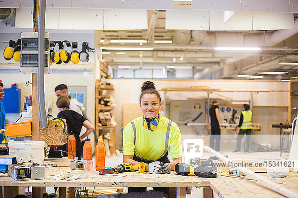 Portrait of smiling young female trainee leaning on workbench with power tools at illuminated workshop