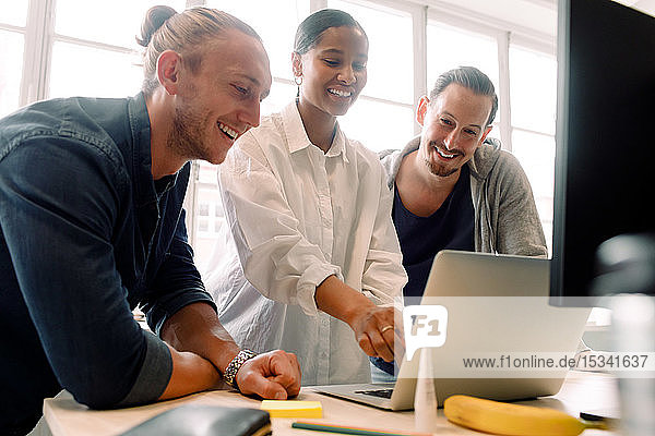 Lächelnde Kollegen schauen auf den Laptop  während sie im Büro diskutieren