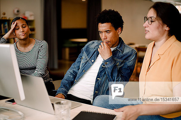 Nachdenklicher Geschäftsmann schaut auf Laptop  während er mit Kolleginnen im Büro sitzt