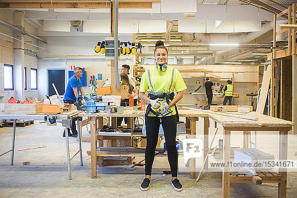 Full length portrait of smiling young female trainee holding power tool while standing by workbench at illuminated works