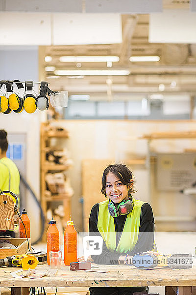 Porträt einer lächelnden jungen Auszubildenden  die sich beim Workshop an die Werkbank lehnt