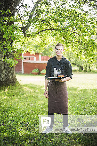 Full length portrait of smiling confident young waiter standing on grass at outdoor cafe