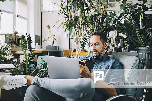 Confident creative businessman using mobile phone while sitting with laptop on seat in office