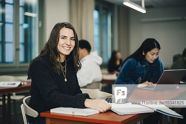 Portrait of smiling teenage girl sitting at desk with classmates in background