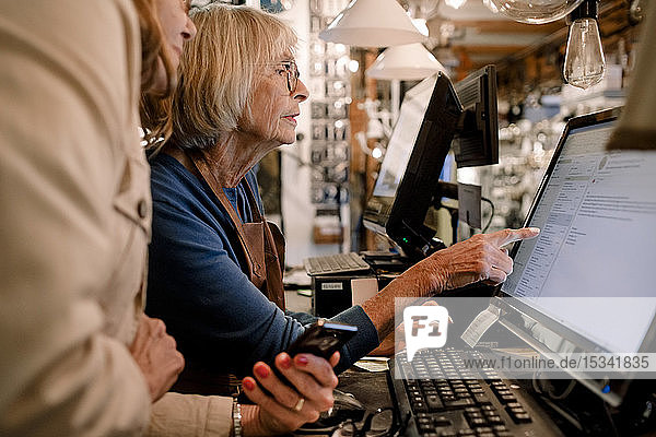 Ältere Kundin und Verkäuferin diskutieren am Computerbildschirm an der Kasse im Baumarkt