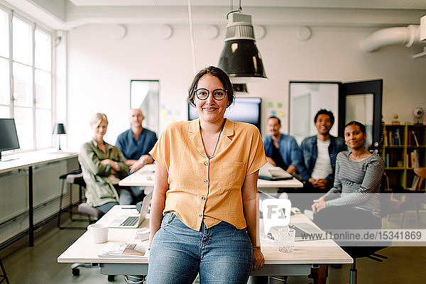Porträt einer lächelnden Berufstätigen  die sich auf dem Tisch gegen Führungskräfte in einem modernen Büro lehnt