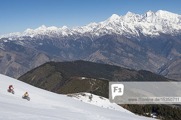 Mountain bikers cycle along a snow covered slope in the Himalayas with views of the Langtang range in the distance  Nepal  Asia