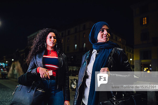 Junge Frau im Hidschab und beste Freundin nachts auf dem Bürgersteig der Stadt