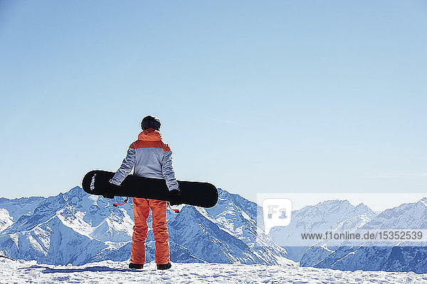 Teenage boy snowboarder looking out over landscape from snow covered mountain top  rear view  Alpe-d'Huez  Rhone-Alpes  France
