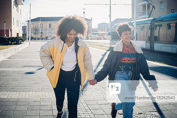 Two young female friends holding hands while strolling on urban sidewalk