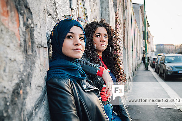 Junge Frau im Hidschab und beste Freundin lehnen an der Wand auf dem Bürgersteig der Stadt