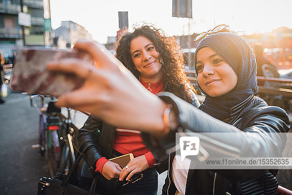 Young woman in hijab with friend taking smartphone selfie in city