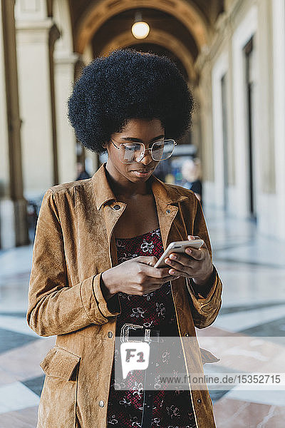Junge Frau mit Afro-Haaren benutzt Smartphone im Gebäudekorridor