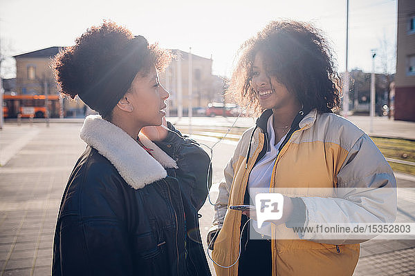 Zwei coole junge Freundinnen teilen sich Kopfhörer auf dem städtischen Bürgersteig