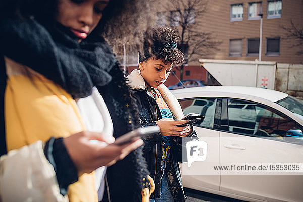 Zwei coole junge Frauen  die auf dem städtischen Bürgersteig spazieren gehen und auf Smartphones schauen  Seitenansicht