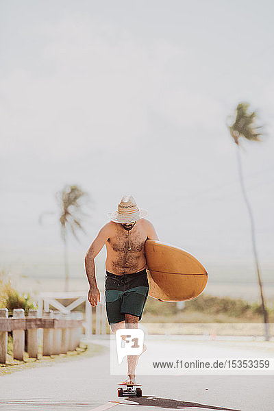 Mittelgroßer erwachsener männlicher Skateboarder mit Surfbrett  Skateboarding auf Küstenstraße  Haiku  Hawaii  USA