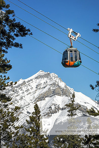 Gondola pod travelling along cables with snow covered mountain and blue sky in the background framed by evergreen trees Banff National Park; Banff Alberta Canada