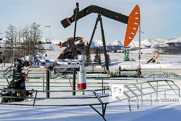 Well head with pipeline and pumpjack in a snow-covered field with snow-covered mountains and blue sky in the background  North of Longview; Alberta  Canada