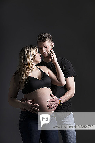 Ein junges,  schwangeres Paar schaut sich an,  und der Vater hält ihren Bauch in einem Studio vor einem dunklen Hintergrund: Edmonton,  Alberta,  Kanada