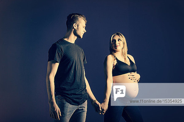 A young expectant couple holding hands with the mother looking at the camera in a studio on a black background: Edmonton  Alberta  Canada