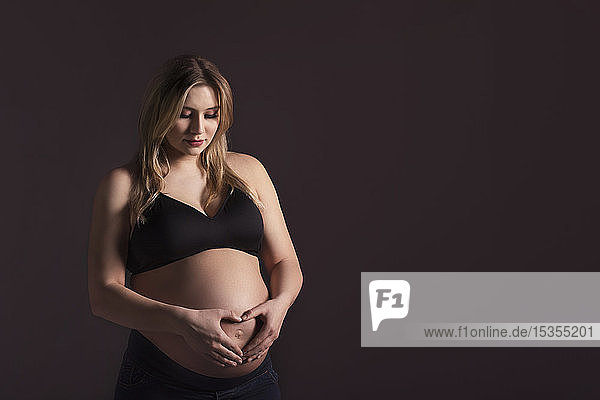 A young pregnant woman holding her belly in a studio and looking down while making a heart shape over her unborn child; Edmonton  Alberta  Canada