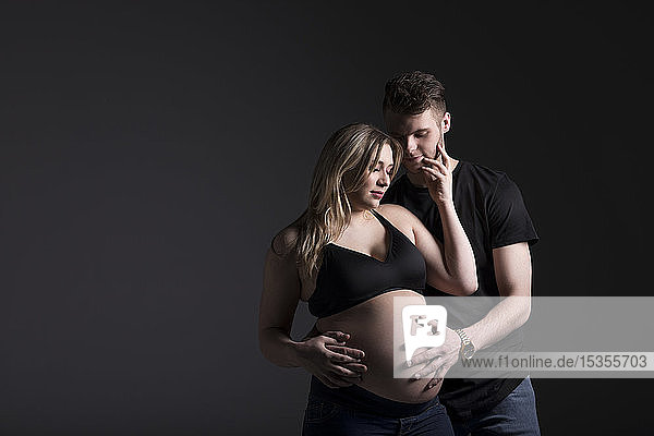 Ein junges schwangeres Paar  das auf sein Baby herabblickt  während der Vater ihren Bauch in einem Studio vor einem dunklen Hintergrund hält: Edmonton  Alberta  Kanada