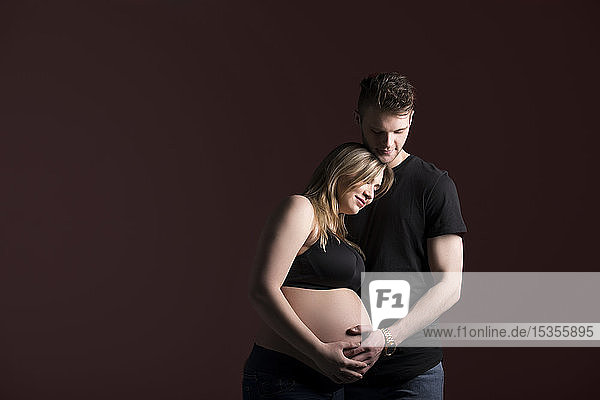 Ein junges schwangeres Paar  das auf sein Baby herabblickt  während es seinen Bauch in einem Studio vor einem schwarzen Hintergrund hält: Edmonton  Alberta  Kanada