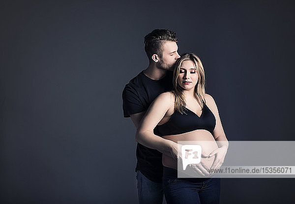 A young expectant couple looking at each other and the father is kissing her forehead and holding her belly in a studio on a dark background; Edmonton  Alberta  Canada