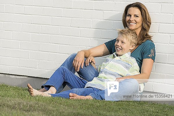 Mutter und Sohn sitzen zusammen vor einer weißen Wand  Mutter schaut in die Kamera  Sohn schaut in die Ferne  Lächeln  Deutschland  Europa