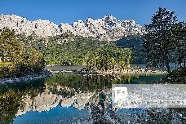 Hiker standing on rocks  view into the camera  Zugspitze and Wetterstein range with reflection in the Eibsee lake  near Grainau  Upper Bavaria  Bavaria  Germany  Europe