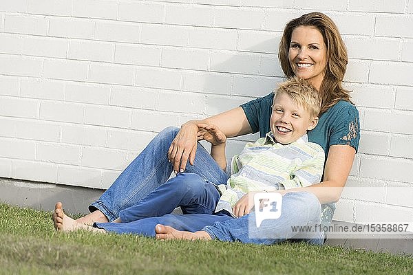 Mutter und Sohn sitzen gemeinsam vor einer weißen Wand  schauen in die Kamera  lächeln  Deutschland  Europa