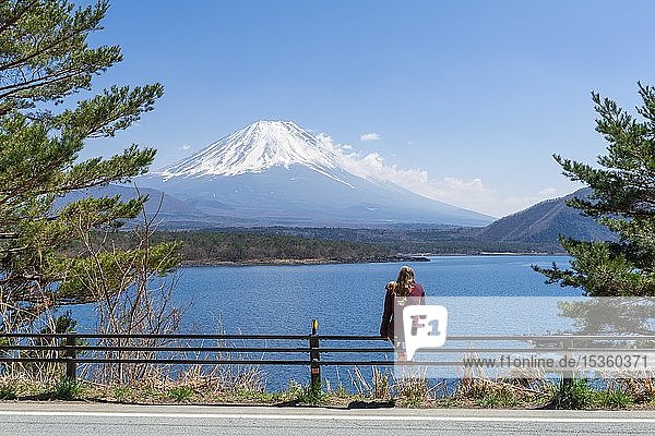 Young woman sitting on a railing next to a road and looking across the lake to the volcano Mt Fuji  Motosu Lake  Yamanashi Prefecture  Japan  Asia