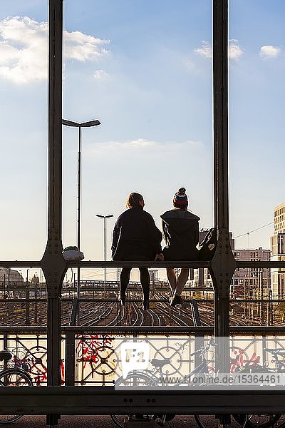 Zwei junge Frauen sitzen auf der Brüstung der Hackerbrücke über die Bahngleise und schauen in die Ferne  Gegenlicht  München  Oberbayern  Bayern  Deutschland  Europa
