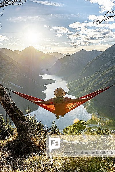 Frau mit Sonnenhut sitzt in einer orangen orangen Hängematte Panoramablick auf Berge mit See Plansee Tirol Österreich Europa