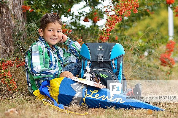 Junge  6 Jahre  erster Schultag  sitzt und lächelt mit Schultasche und Schultüte am Baum  Deutschland  Europa