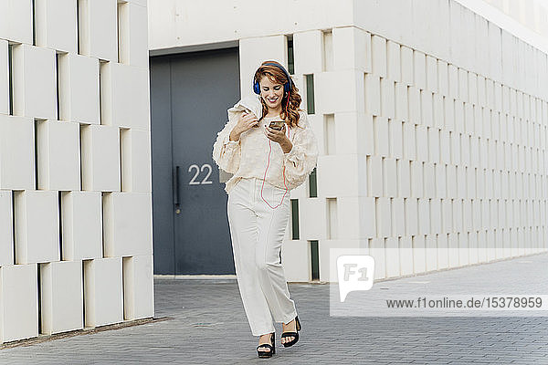 Geschäftsfrau geht auf die Straße und hört mit Kopfhörern Musik von ihrem Smartphone