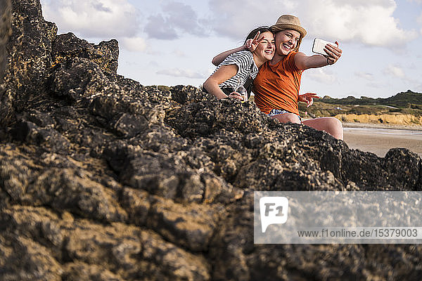 Two girlfriends sitting on rocky beach  taking smartphone selfies
