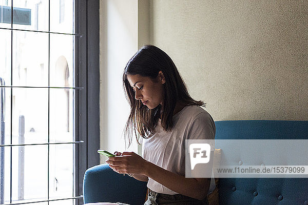 Junge Frau sitzt auf einer Couch am Fenster und benutzt ihr Handy