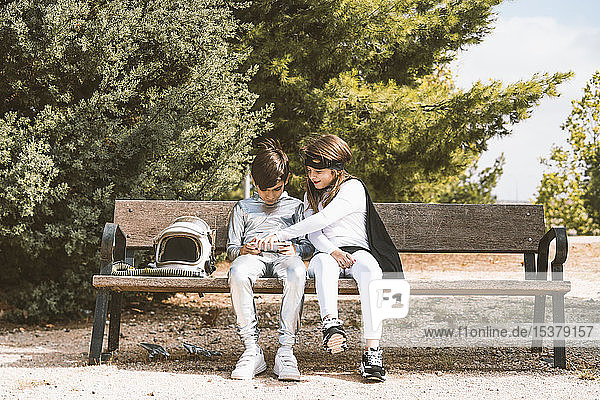 Two kids in astronaut and superhero costumes using mobile phone on park bench